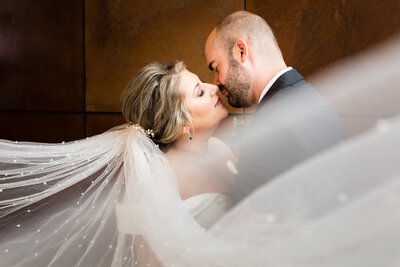 A bride's veil sweeps in front of an almost kissing newlywed couple in front of the large copper plated wall in Avenue which is located in Greenville SC.
