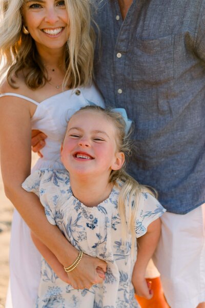 A young girl in a blue and white dress smiles big while posing on the beach for family photos in Los Angeles with her mom and dad.