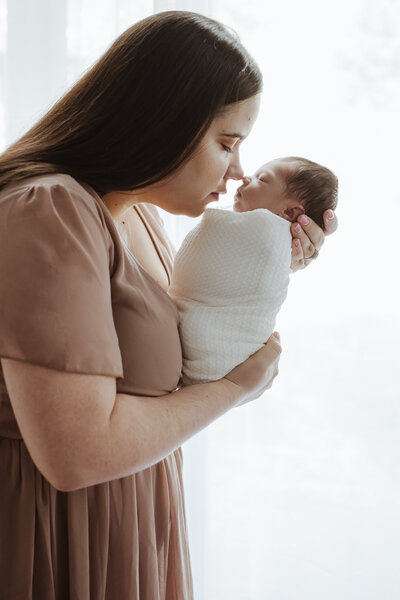 Motherhood photo of newborn baby girl wrapped in white blanket being held by mom in pink dress and touching noses.