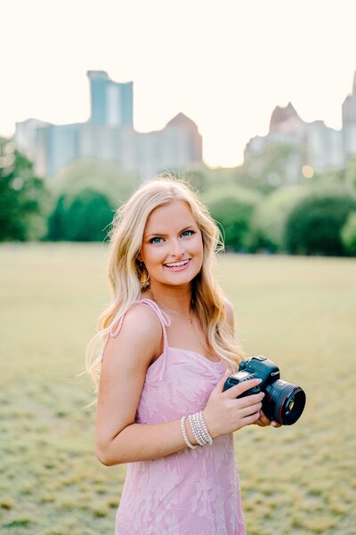 A branding photo for a woman wedding videographer and woman photographer holding a camera in a field in the city.