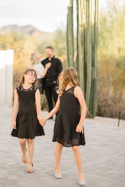 two girls walking holding hands with mom and dad in the background smiling at each other 