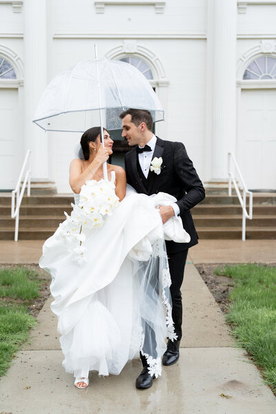 The couple is looking at each other as they walk in the the rain. Bride is holding an umbrella as groom holds her dress. 