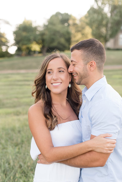 Engaged couple hugging tightly as guy gently rests his nose on girl's left temple on sunny evening.