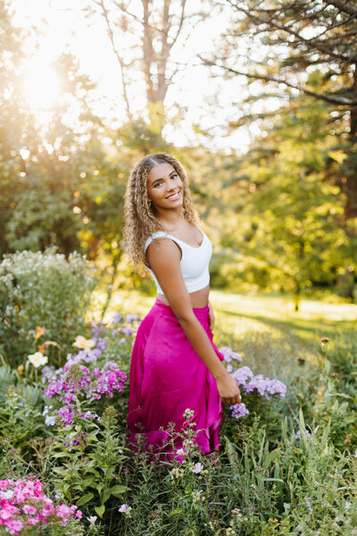 Senior girl in a white top and pink skirt twirling among flowers and tall grass with the sun setting in the background