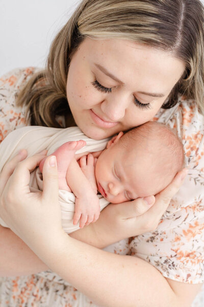 New Mom in beige floral dress with professional hair and makeup done. She is holding her newborn baby up by her face and has her eyes closed. Baby is also sleeping and wrapped in a beige swaddle his feet and hands are showing.