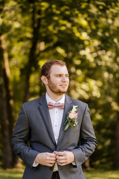 Groom in tux wearing a soft pink bow tie looking away and buttoning jacket wedding at Barn at Murphey Farm