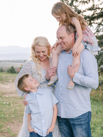 Family of four snuggling and smiling at each other with their young daughter on the dads shoulders all dressed in nice blue toned colors-  Maegan R Photography