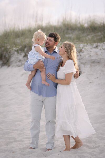 A young family shares a joyful moment during their beach photo session at Neptune Beach, Florida. The father holds their blonde toddler while the mother, dressed in a flowing white dress, smiles up at them. Barefoot in the sand and framed by soft dunes and natural grasses, the scene feels warm, candid, and timeless. Captured by a Jacksonville family photographer specializing in relaxed, natural beach portraits that celebrate connection and love.