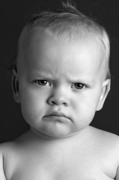 Black and white close-up of a baby with a furrowed brow, giving a stern expression. The mood is serious, with a neutral, dark background.