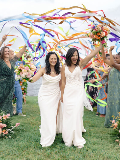 Two brides in white dresses joyfully walk hand in hand, surrounded by cheering guests waving colorful ribbons. Each bride holds a bouquet of flowers. The scene is vibrant and celebratory, set outdoors on a grassy area.