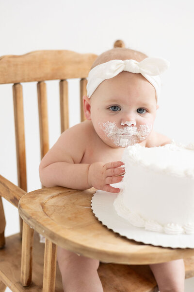 One year old girl with a white bow on her head eats a large cake during her cake smash session at Kathleen Jablonski Photography studio.