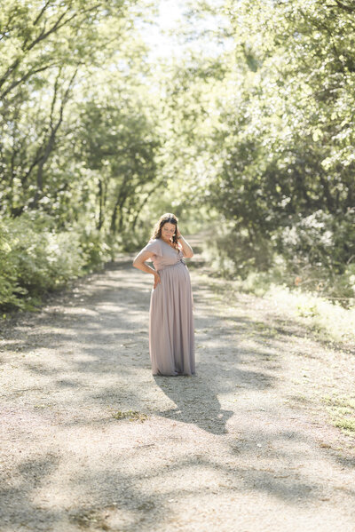 Pregnant woman standing in soft sunlight on a wooded trail, wearing a blush dress during a Kansas City maternity session.