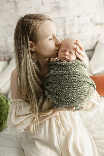 A serene family portrait featuring a mother and father seated together, lovingly holding their newborn baby. The baby is peacefully swaddled in a cozy green knit blanket. The mother wears a soft, off-the-shoulder cream dress, and the father, in a casual cream shirt and glasses, gazes at the baby with affection. The neutral tones and textured backdrop add warmth and intimacy to the image.