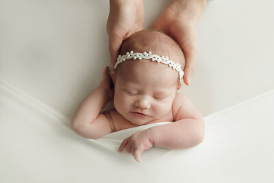 Newborn baby sleeps with her parents hands behind her head.