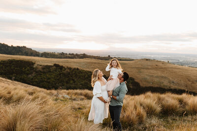 A father holds his daughter on his shoulders while the mother looks on during a sunset family photoshoot in the hills in christchurch