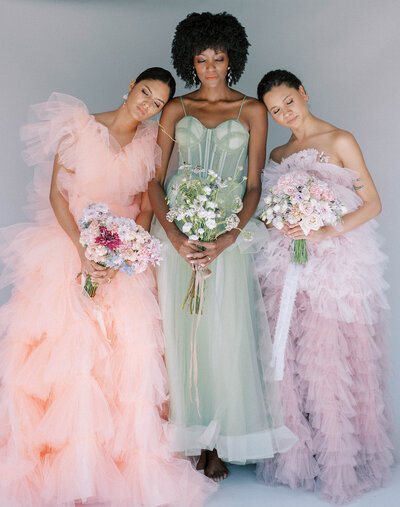 group of three bridal models standing together while holding bouquets of florals, each in a different color gown