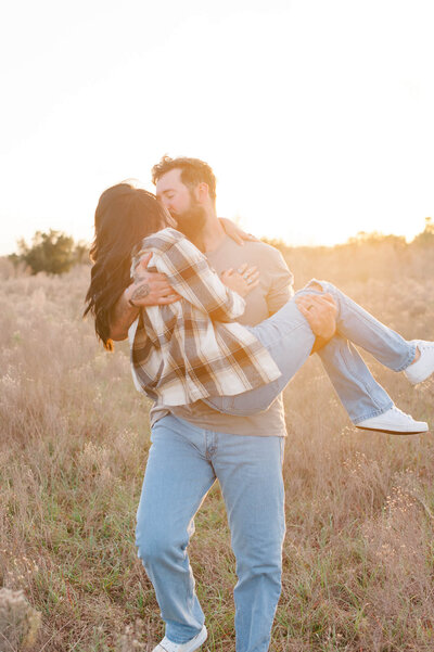 Couple kissing while he holds her through a tall grass field at sunset with a beautiful golden hour glow