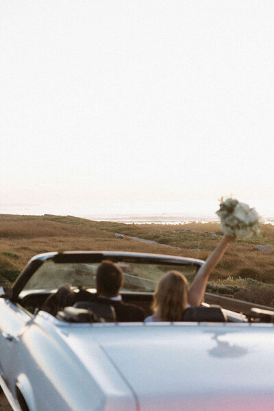Coastal vintage inspired engagement session. Couple in vintage car driving down the coast in a vintage blue convertable while bride holds wedding bouquet in the air