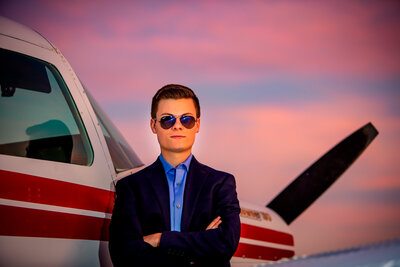 A high school senior boy wearing a suit & sunglasses, posing in front of a plane & sunset sky for a south shore ma senior photographer