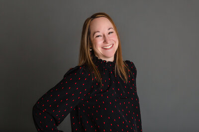 Woman with medium, copper-colored hair smiles at camera wearing a black blouse with red dots.