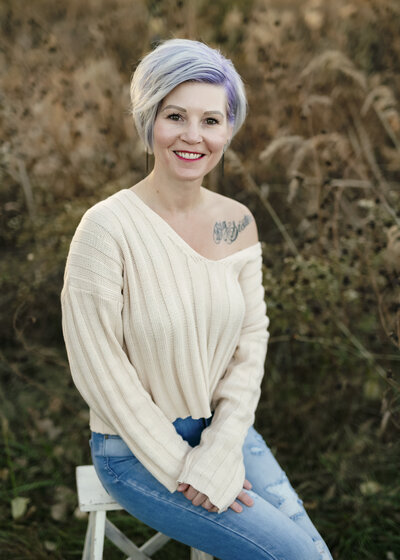 Brieanna Lebo sitting on a wooden chair outdoors in a cream sweater and ripped jeans, natural fall setting.