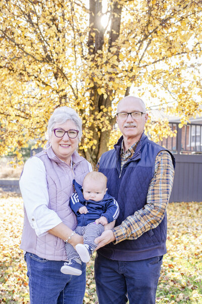 Grandparents holding grandbaby boy in front of a yellow leafed tree on a Fall afternoon in Mt. Orab, Ohio.