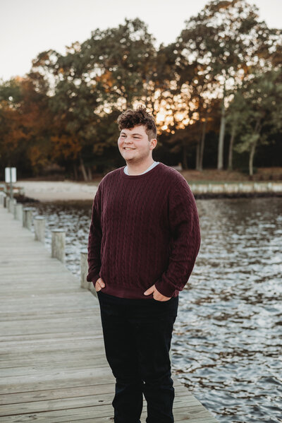Senior boy wearing red sweater and black pants smiling standing on a pier with sun shining behind him.