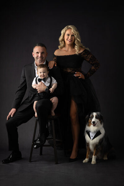 A family portrait captured by a Dallas family photographer, featuring a couple with their three children. The family wears coordinated gold-toned dresses and outfits. The mother holds her youngest child, while the father sits with their older children. The photo is set against a rich, dark background that complements the warm tones of their clothing.