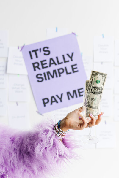 Hand holding a glass filled with dollar bills in front of a purple sign that says, It's really simple pay me.