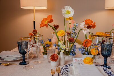Sophisticated wedding tablescape with bright orange and yellow flowers, blue goblets, and gold-accented tableware for a refined event setting.