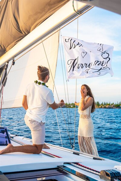Woman reacting with joy as her partner proposes on a sailboat in Hawaii, standing beside a waving Marry Me flag over the ocean.
