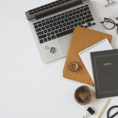 An arial view of a desk displaying an open laptop, a closed iPad with a gold cover, two notebooks, glasses and paperclips are displayed on a white surface.