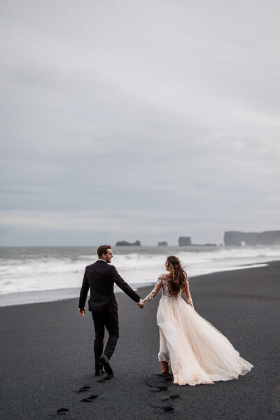 A bride and groom walk on a black sand beach in Iceland