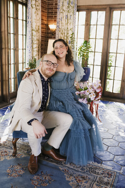 Bridge wearing blue tulle dress with bouquet sits on groom's lap indoors at vintage chic venue.