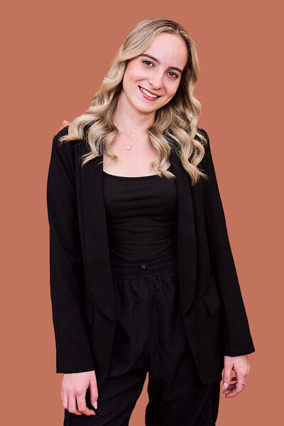 Bailey, Male hairdresser with styled short hair, dressed in a relaxed black outfit against a warm-toned backdrop.