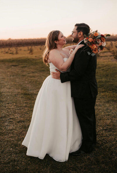 bride holding a wedding bouquet wearing a simple white gown and a beaded headband, smiling at the groom who is dressed in a black suit, as he gently holds her