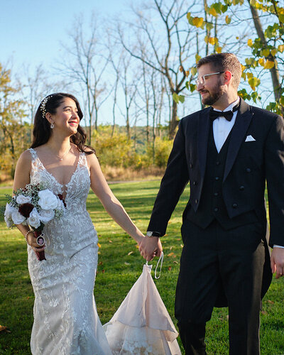 A bride in a white lace gown holds a bouquet and smiles at a groom in a black tuxedo as they walk hand in hand on a grassy lawn, with trees and sunlight in the background—captured by an experienced nj wedding photographer.