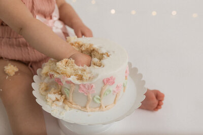 Close-up of a baby’s hand smashing into a decorated cake with pink flowers during a cake smash photoshoot.