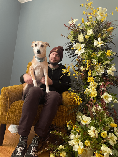 Man sitting on couch with dog on his lap and yellow themed floral display next to them. Designed by Jessamine floral and events, New Jersey floral designer