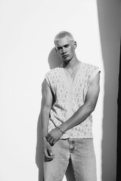 Black and white editorial portrait of a young man with platinum blond hair, wearing a textured sleeveless sweater. Shot in dramatic lighting for a bold, masculine fashion look.