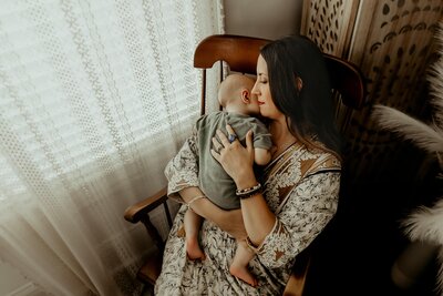 In a warm and cozy studio setting, a new mother lovingly cradles her baby in her hands while sitting in a boho peacock chair. With a mixture of awe and tenderness, she gazes down at her precious little one. This heartfelt moment is artfully captured by Fire Family Photography, a newborn photographer from Macon, GA.