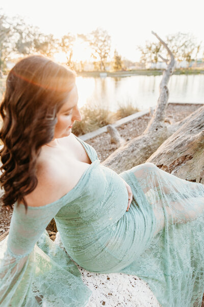 pregnant mom sitting on fallen tree trunk and holding baby bump at sunset