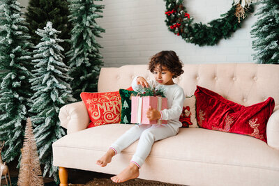 Wandermama Photography children mini session with christmas decoration around him. He is wearing pajama and in his legs are a gift. He is sitting on the white couch decorated with red and green cushions