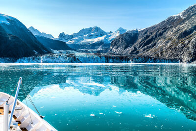 A scenic view of a glacier surrounded by snow-covered mountains, reflected in the calm turquoise water, with the edge of a boat visible in the foreground.