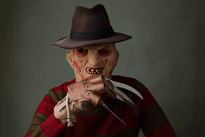 Male dressed up in Freddy Krueger costume with claw underneath his chin sitting in front of dark green backdrop.