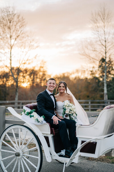 man in black tux and woman in white dress sitting in white carriage in front of the sunset