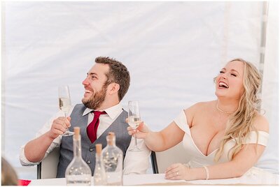 bride and groom laughing during the speeches while holding champagne glasses and sitting down at the reception table