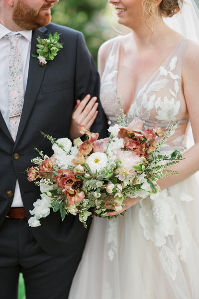 bride loops her arm through the grooms who is wearing a navy blue suit and floral tie bride is wearing a grey wedding dress with a floral overlay groom has an all greenery boutonniere and bride holds her bridal bouquet made by cincinnati wedding florist roots floral design