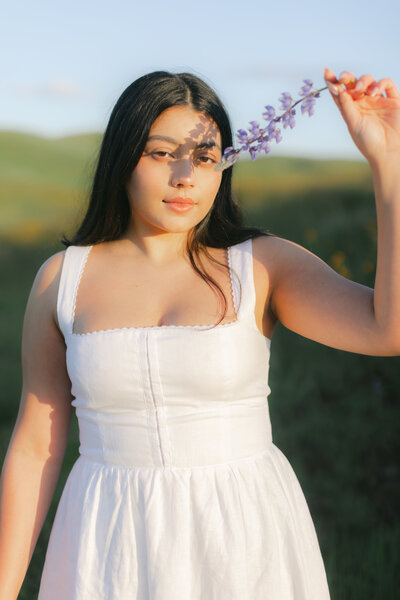 Editorial portrait of a young girl in a white dress holding a purple flower over her eye, casting a soft shadow on her face during springtime in Fresno, California.
