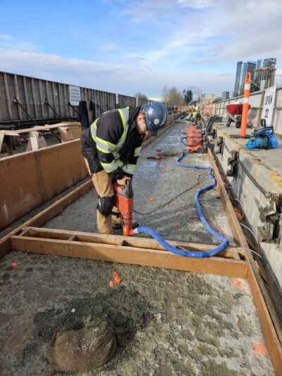 An operator on a site operating heavy machinery to drill holes into existing concrete and inserting steel dowels to transfer loads and prevent cracking at joints.